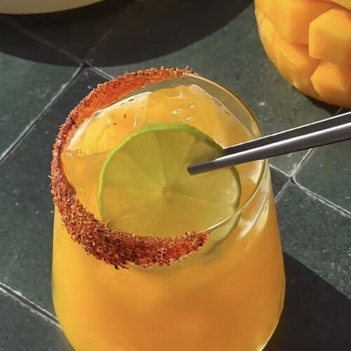 Close-up of a chamoy margarita with a chili-salt rim, lime garnish, and metal straw on a dark marble surface